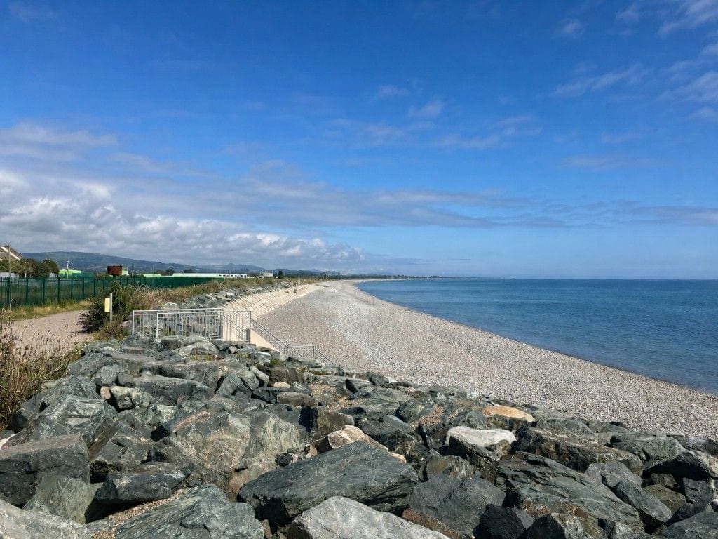Shingle behind rock armour, steps and railings; a green palisade and industrial buildings inland.